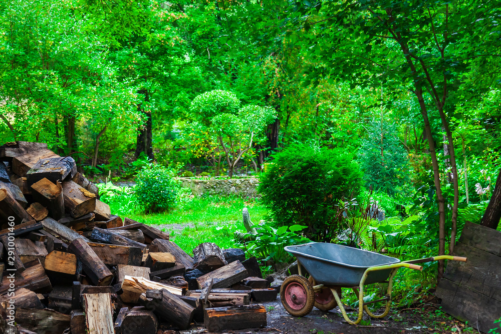 A pile of firewood in the yard near a wooden house made of timber and a cart nearby in a picturesque place, for kindling a stove or bath.
