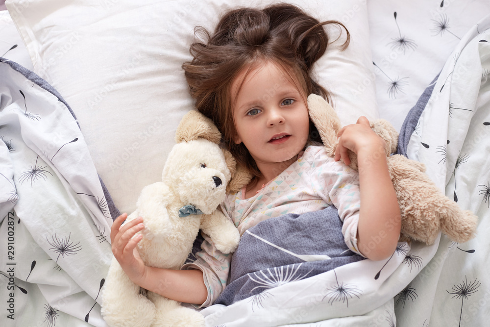 Horizontal shot of toddler girl in bed lying with his teddy bear at home, sleeppy child laying on linens with dandelion, looking directly at camera, has bcalm facial expression. Childhood concept. Horizontal shot of toddler girl in bed lying with his teddy bear at home, sleeppy child laying on linens with dandelion, looking directly at camera, has bcalm facial expression. Childhood concept.