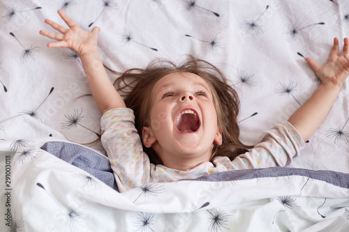 Close up portrait of cheerful little girl lying in bed under blanket with dandelion, charming kid wearing pajama, spreading her arms, keeps mouth openes, screams something happily. Childhood concept. photo
