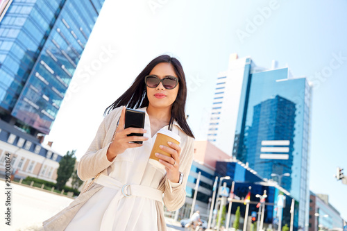 communication, lifestyle and technology concept - happy smiling young asian woman in sunglasses with takeaway coffee cup and smartphone on city street