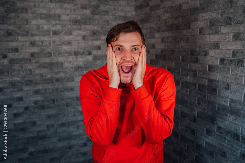 A beautiful emotional man in a red hoodie is happily surprised with hands on her cheeks. A beautiful emotional man in a red hoodie is happily surprised with hands on her cheeks.