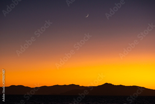 Fotografija Glistening new moon sickle setting behind the dark hills against an orange sunset, with water in the foreground