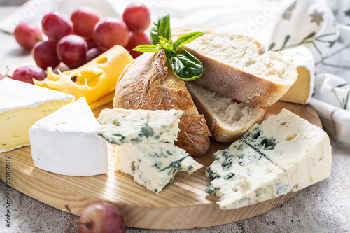 Assortment of cheese with ripe red grapes , aromatic basil and French baguette on the wooden tray close up. Healthy and tasty food concept.