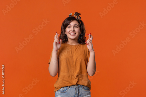 Smiling brunette woman in an orange t-shirt and beautiful headband holding fingers crossed asking for a dream come true standing isolated over orange background. Concept of dreaming.