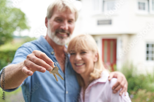 Portrait Of Mature Couple Standing In Garden In Front Of Dream Home In Countryside Holding Keys