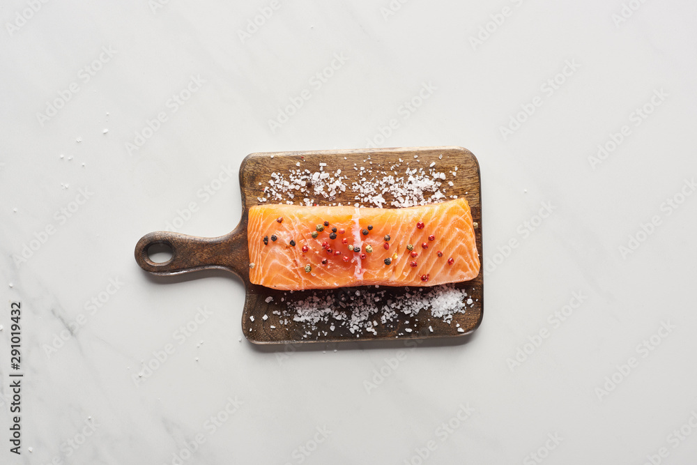 top view of raw salmon steak with peppercorns and salt on wooden cutting board on marble surface