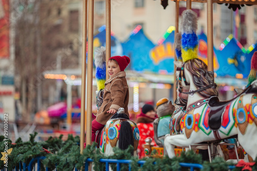 Cute smiling boy in a brown coat and a red hat rides on a merry-go-round.