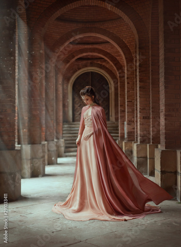 Sad princess in a vintage royal nude powdery color dress and in a cloak that flies in the wind. The magical Rays of the sun and hope pour on a woman through the columns. Long dark hair with a tiara.