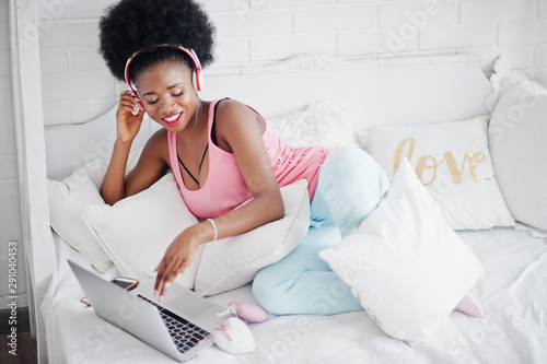 Young african american woman sitting in bed while working on laptop and listen music on earphones.