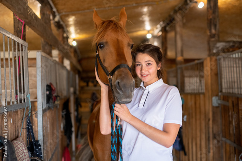 Pretty young smiling brunette keeping one hand by muzzle of purebred brown mare