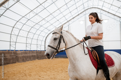 Serious young active woman looking straight while riding white purebred horse