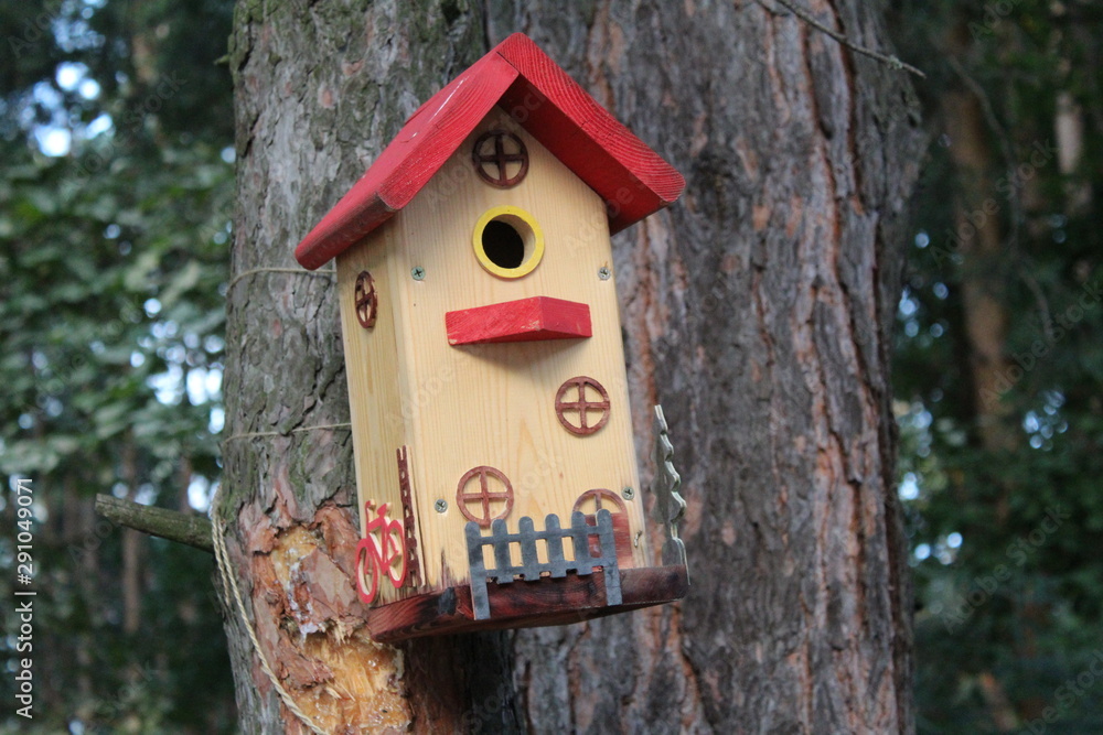photo of the birdhouse for the birds in the Park.house made with your own hands.the material of wooden.the house is painted a yellow color.on it jewelry.time of year summer.trees with green foliage.