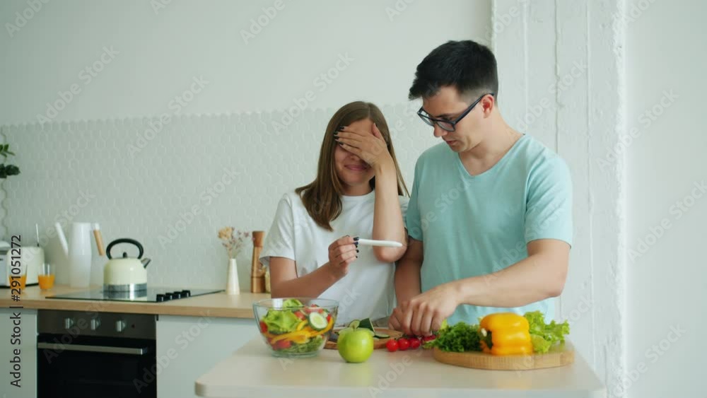 Man husband is cooking food in kitchen cutting salad when happy girl is bringing pregnancy test, people are talking smiling. Family and modern lifestyle concept. Man husband is cooking food in kitchen cutting salad when happy girl is bringing pregnancy test, people are talking smiling. Family and modern lifestyle concept.
