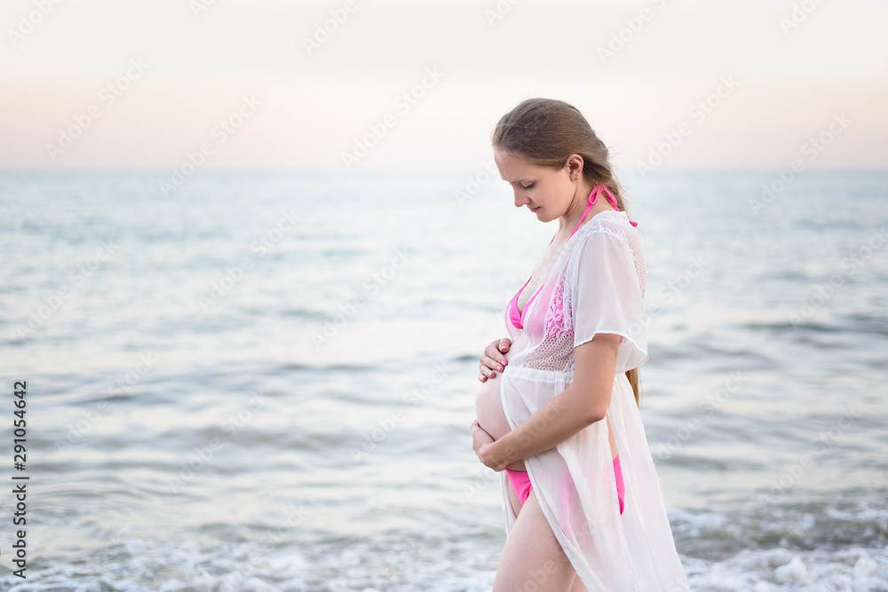 Young pregnant woman is standing on the seashore and hugging her belly. Enjoying the moment. Young pregnant woman is standing on the seashore and hugging her belly. Enjoying the moment.