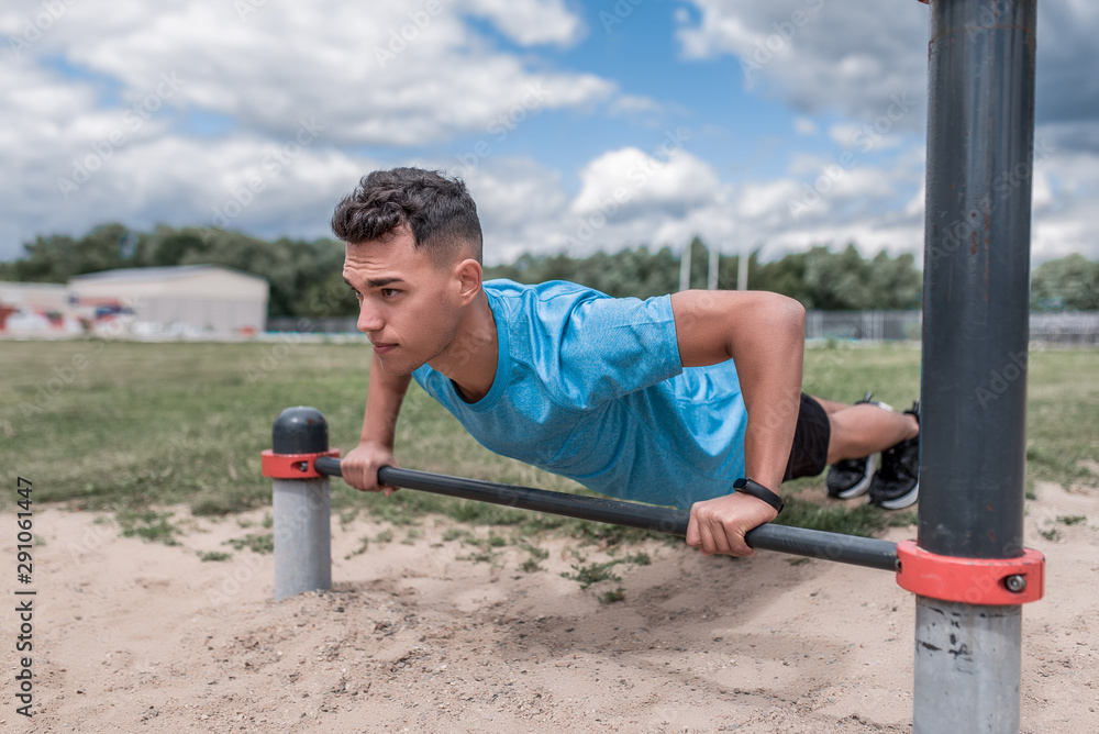 male athlete is pushing up from horizontal bar, training is powerful, in summer in city, in afternoon in fresh air. Youth lifestyle, active modern guy athlete. Background grass sand sky.