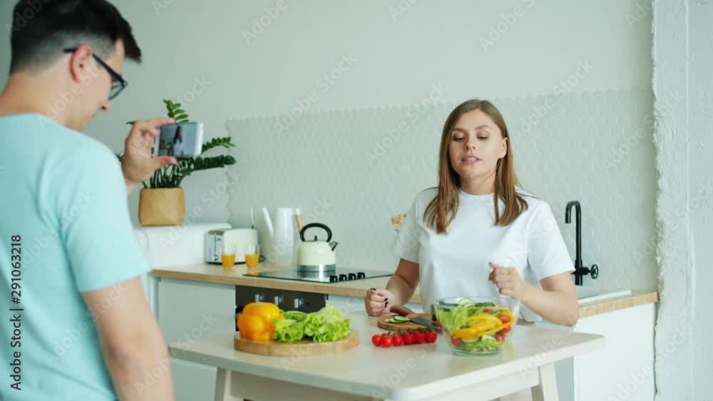 Family of vloggers man and woman are recording video in kitchen talking cooking food holding vegetables using smartphone camera. Vlogging and people concept. Family of vloggers man and woman are recording video in kitchen talking cooking food holding vegetables using smartphone camera. Vlogging and people concept.