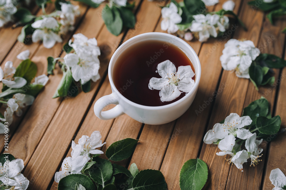 White cup of tea with petals of an apple tree on a wooden background, view from above, close up.