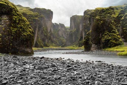 Entrance of a large icelandic canyon with a torrent flowing in the middle, surrounded by pebbles
