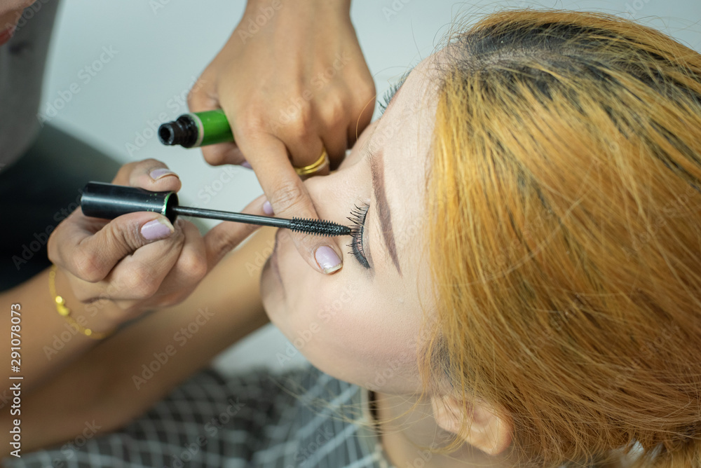 Makeup artist asian woman applying cosmetic mascara on eyelashes using curling brush.