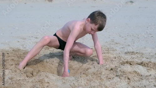 Child boy playing on sand beach digging shovel deep hole and throwing sand on sea ocean coast. Outdoor summer activities at vacation. Boy is sunbathing in swimming trunks.