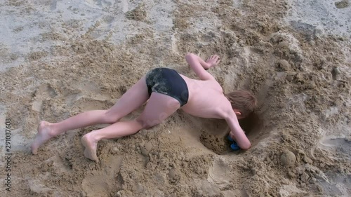 Child boy playing on sand beach digging shovel deep hole on summer vacation on sea ocean coast, top view. Outdoor summer activities on vacation. Boy is sunbathing in swimming trunks.
