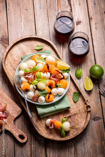 Melon, mozzarella, prosciutto appetizer or snack, cantaloupe melon, top view, rustic wooden background, skewer, party food, two glasses of wine