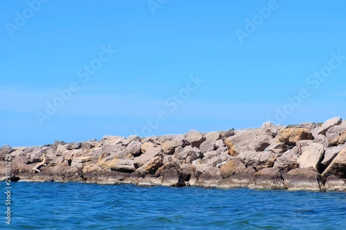 Wild seashore with stones, blue sky and water at sunny day. Nature background seascape. Beautiful natural coastline place. Pier from cobblestones at edges.