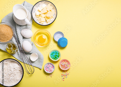 Baking ingredients for cake, cupcakes or muffins on yellow copy space background for text, sugar pearls, eggs, whisk, flour, butter, sugar and milk