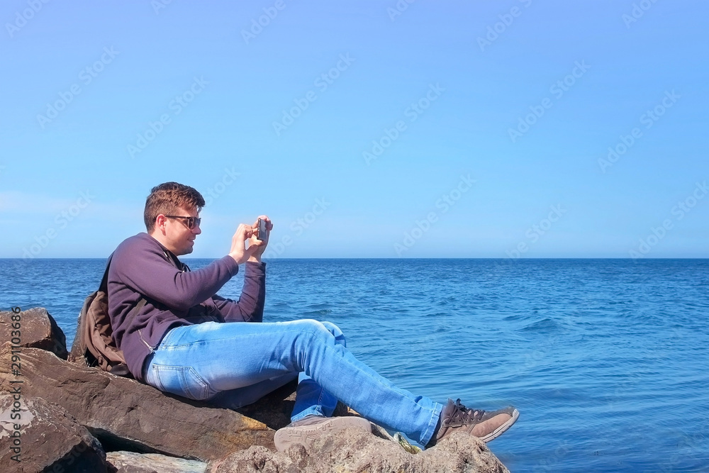 Young man tourist traveller taking photos on smartphone sits on boulder at sea. Guy blogger working photographing beautiful seascape on mobile phone. Tourism travel journey vacation on ocean concept.