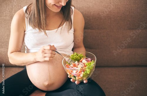 Front view of pregnant woman sitting on sofa at home and eating fresh salad. Happy mother eating vegetables and caring about health and baby. Concept of expectation and health.