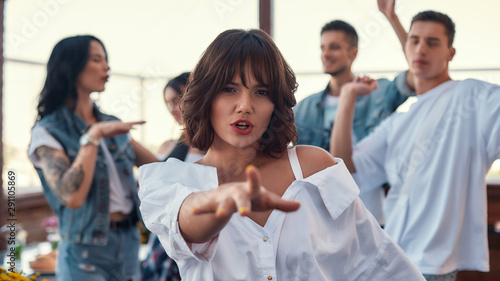 Dancing with friends. Pretty and young woman in white shirt is gesturing and looking at camera while standing on rooftop terrace with her friends