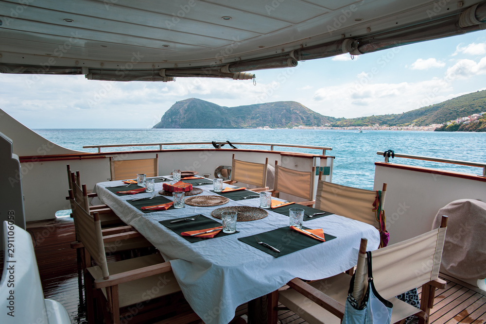 Naklejka premium Table set for breakfast on the rear deck of a luxury private yacht, cruising the mediterranean sea and docked in front of Lipari Island.