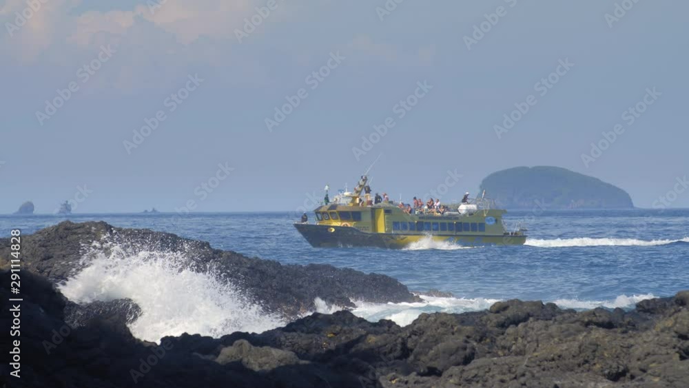 Small boat with tourists is passing green navigational buoy on big blue waves near black volcanic rock with huge splashes and huge oil tanker ship in background Small boat with tourists is passing green navigational buoy on big blue waves near black volcanic rock with huge splashes and huge oil tanker ship in background
