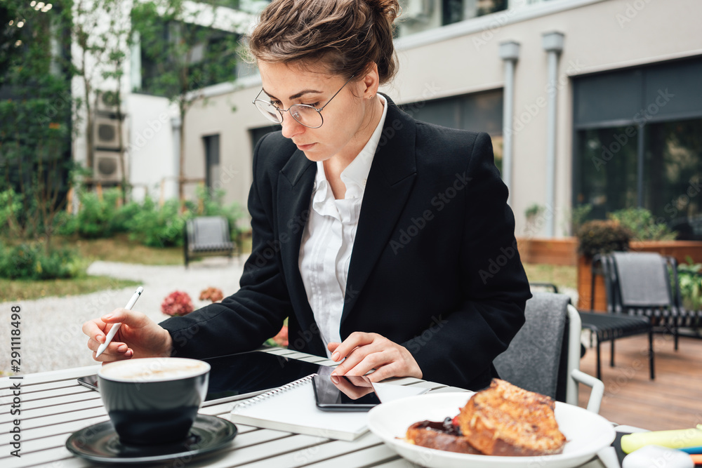 Portrait young businesswoman sitting in cafe on veranda and with a digital tablet. Pensive woman wearing glasses using tablet for notes Portrait young businesswoman sitting in cafe on veranda and with a digital tablet. Pensive woman wearing glasses using tablet for notes
