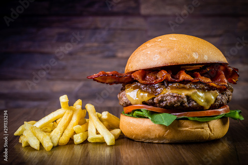 Fotografie Craft beef burger with cheese, bacon, rocket leafs, caramelize onion and french fries on wood table and rustic background
