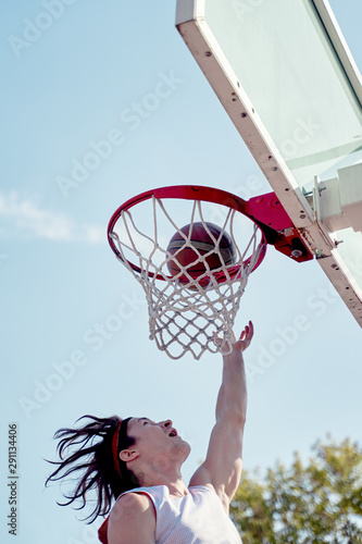 Photo of young athlete man throwing ball into basketball hoop on sports field on street on summer day. © Sergey