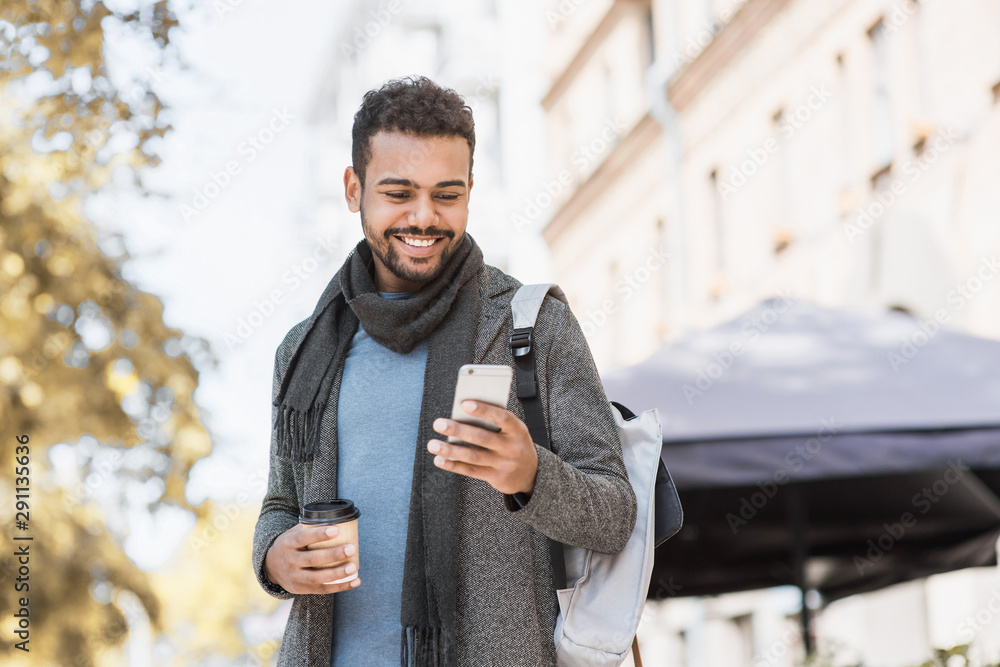 Fototapeta premium Young handsome student man using smartphone. Smiling joyful guy autumn portrait. Cheerful men wearing warm clothes holding mobile phone in a city in winter. 