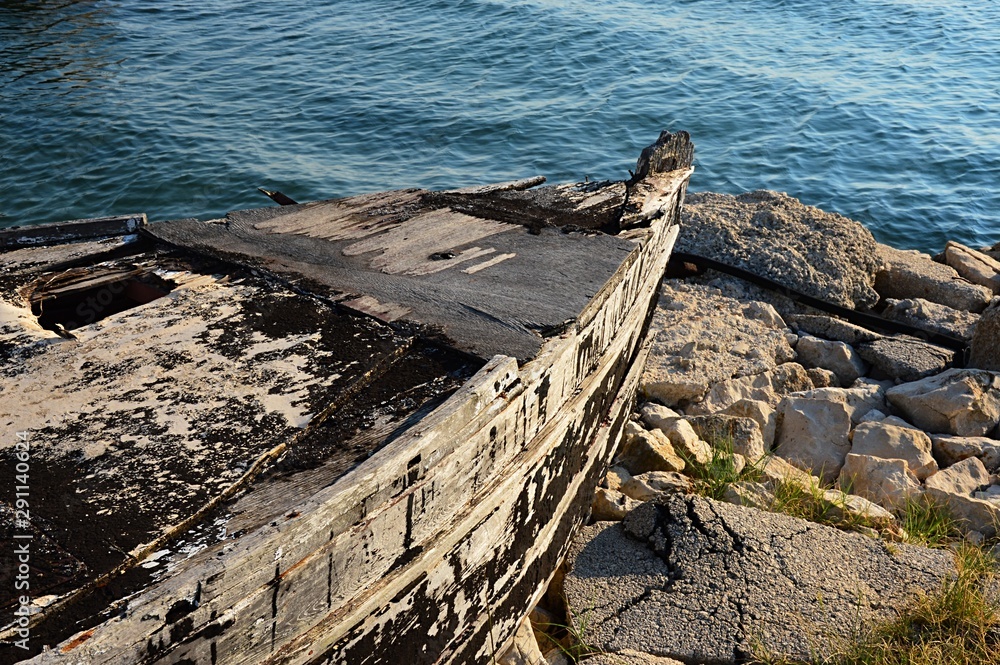 Front part of old decaying fishing boat placed on damaged rocky molo, used as storage for fishnets and fishing equipment, small waves on sea water surface visible Front part of old decaying fishing boat placed on damaged rocky molo, used as storage for fishnets and fishing equipment, small waves on sea water surface visible