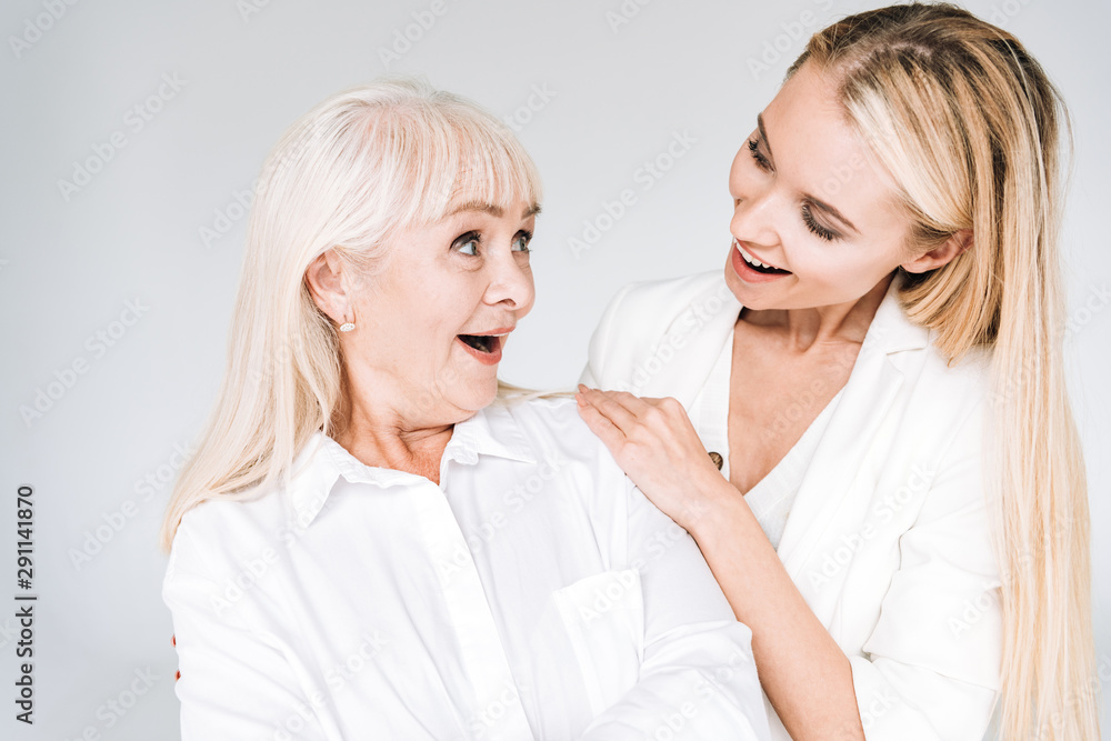 excited blonde grandmother and granddaughter together in total white outfits looking at each other isolated on grey excited blonde grandmother and granddaughter together in total white outfits looking at each other isolated on grey