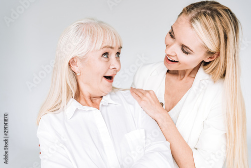 excited blonde grandmother and granddaughter together in total white outfits looking at each other isolated on grey