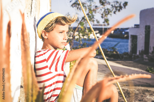 Portrait of a young fisherman sitting near the harbour with fishing pole looking at the sea. Sailor boy with wool cap and a striped t-shirt relaxing in the evening sun. Lifestyle outdoor concept