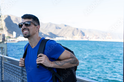 Happy fit traveler with sunglasses and tattoo in the arms smiling in summertime, excited for his journey. Handsome black hair tanned backpacker standing in front of the sea