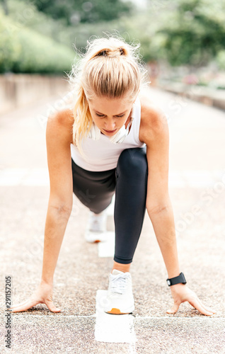 Full-length portrait of motivated woman getting ready to start running on stadium. Young sportswoman with headphones and smartwatch going to run outdoors. Summer training concept. Vertical shot © Adamov
