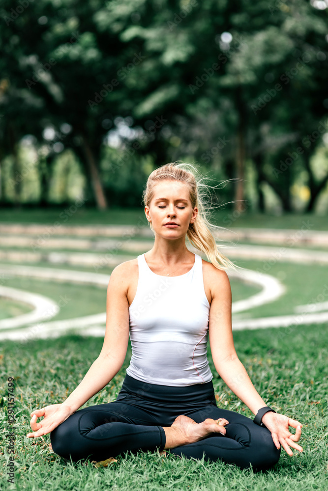Full-length portrait of young caucasian woman in sportswear practicing yoga. She is sitting in Lotus pose in the city park. Outdoors meditation concept. Vertical shot. Front view Full-length portrait of young caucasian woman in sportswear practicing yoga. She is sitting in Lotus pose in the city park. Outdoors meditation concept. Vertical shot. Front view