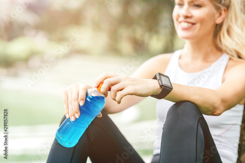 Cropped portait of sporty woman resting after exercising. She is smiling while sitting in the park and holding classic fitness shaker with pre-workout drink. Horizontal shot. Selective focus