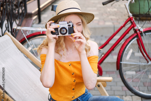 Attractive blond girl in hat dreamily taking photo on retro camera while sitting on deck chair in street cafe © Anton