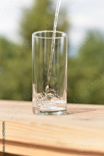 A stream of water flowing into an empty transparent glass standing on a wooden board.
