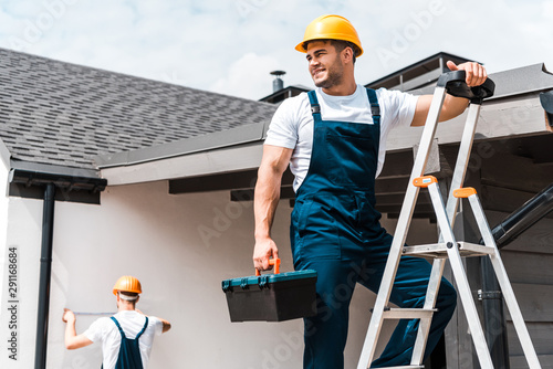 Fotografi selective focus of happy workman standing on ladder with toolbox near coworker
