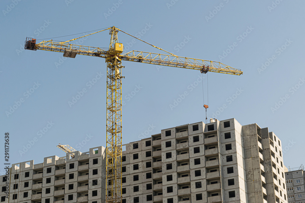 A yellow high-rise building crane against a blue sky builds multi-storey apartment buildings using modern technologies of metal, concrete and brick according to an architectural project A yellow high-rise building crane against a blue sky builds multi-storey apartment buildings using modern technologies of metal, concrete and brick according to an architectural project