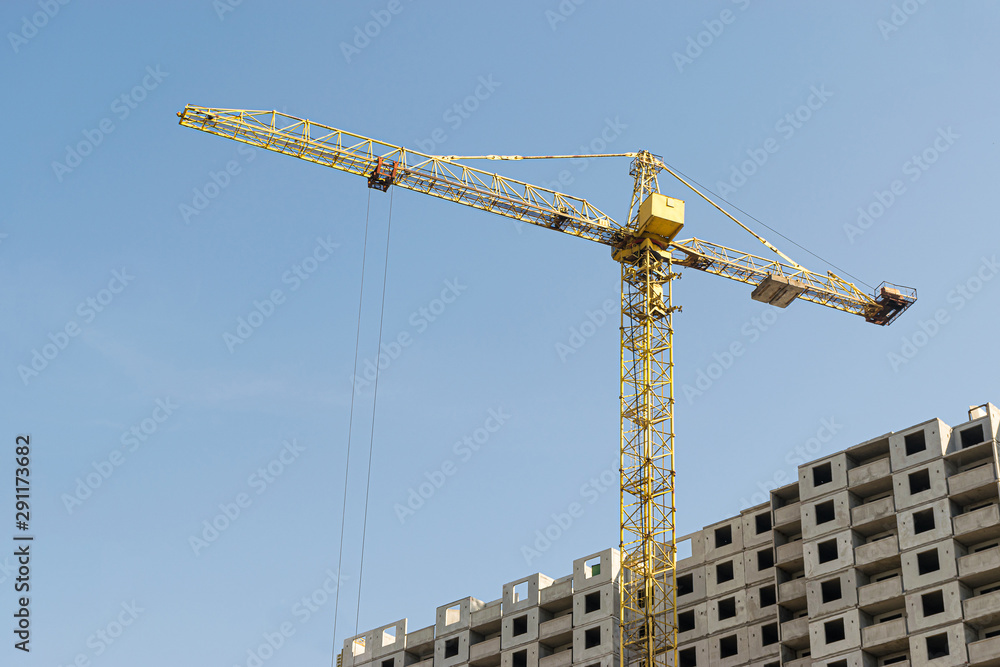 A yellow high-rise building crane against a blue sky builds multi-storey apartment buildings using modern technologies of metal, concrete and brick according to an architectural project